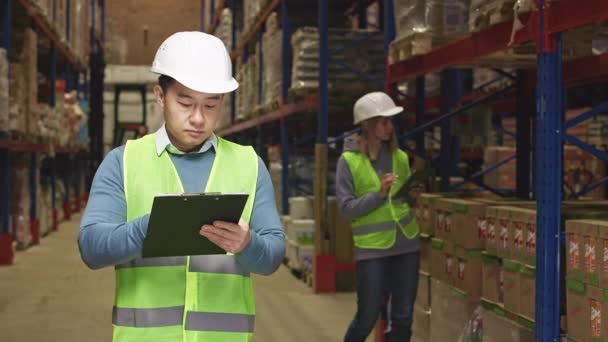 焦点是亚洲男人在剪贴板上记笔记 而高加索女人则站在货架旁边 拿着商品 用数码平板电脑 对工业仓库的视察 — 图库视频影像
