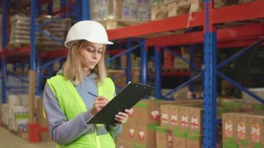 Beyaz kadın rafların arasında elinde paketlerle duruyor, panoya yazıyor ve kameraya bakıyor. Endüstri deposunda beyaz kasketli ve yansıtıcı yelek giyen kadın yönetici..