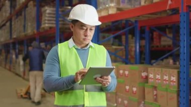 Sanayi deposunda dikilirken dijital tablet üzerinde şirket uygulaması kullanan beyaz kasketli ve yansıtıcı yelekli dikkatli Asyalı bir adam. Üniformalı erkek işçiler arka plana paket yüklüyorlar.