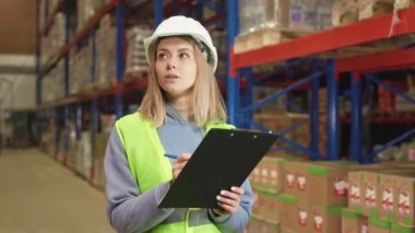 Beyaz saçlı, güvenlik yeleği giyen, beyaz şapkalı bir kadın büyük bir depoda malları sayarken. Fiziksel envanter, lojistik ve yönetim kavramı.