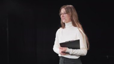 Günlük giysiler içinde gülümseyen beyaz kadın meşgul seyircilere iş seminerlerini dinledikleri için teşekkür ediyor. Sahnede kulaklık ve not panosuyla duran kadın akıl hocası.