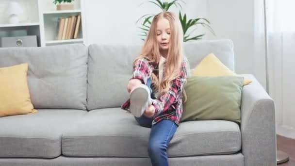 积极的高加索女孩坐在灰色柔软的沙发上 穿着白色的运动鞋 有魅力的孩子穿着休闲装 调整长发 走出家门 — 图库视频影像