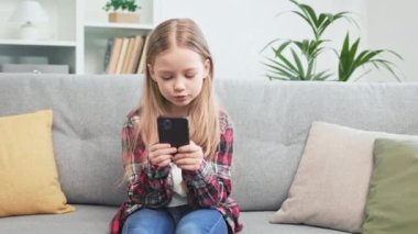 Kafkasyalı altın saçlı kız okul arkadaşlarıyla çevrimiçi sohbet etmek için akıllı telefon kullanarak rahat bir kanepeye yerleşti. İlerici çocuk sürekli iletişim halinde olmak için kablosuz teknoloji kullanıyor..