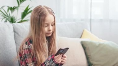 Uzun saçlı sarışın çocuğun rahat bir oturma odasında cep telefonuyla müzik dinlemesinin yan görüntüsü. Sıradan giyimli tatlı okul kızı kablosuz teknoloji kullanarak yeni şarkıların tadını çıkarıyor..