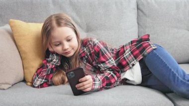 Günlük kıyafetleriyle rahat tatlı kız rahat koltukta yumuşak yastığa uzanıp akıllı telefondan izliyor. İnternette sörf yapan sevimli bir çocuk ve yoğun bir günün ardından dinleniyor..
