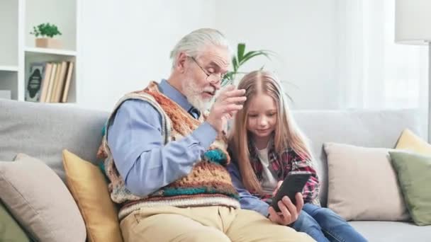 戴眼镜的白人老人和在手机上看滑稽视频的可爱女孩 爷爷和孙女在一起开心地度过了一段快乐的时光 最后享受着与家人团聚的快乐时光 — 图库视频影像