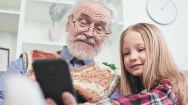 Kaygısız büyükbaba ve torunun kişisel aletlerle ilginç video oyunları oynamaları düşük açılı bir bakış açısı. Küçük çocuk büyükbabasına modern akıllı telefonlarda medya uygulamalarını nasıl kullanacağını gösteriyor..