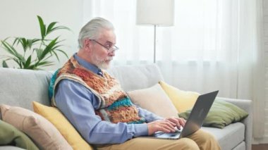 Güzel dedemin rahat oturma odasında kanepede dinlenmesi ve modern taşınabilir dizüstü bilgisayarda internete göz atması. İlerici emekliler emekliliğin keyfini çıkarıyorlar.