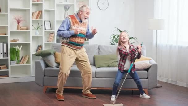 积极的祖父与孙女清洁公寓和跳舞 可爱的小女孩用拖把在地板上擦洗时 这位时髦的老人却很开心 幸福家庭和退休生活的概念 — 图库视频影像