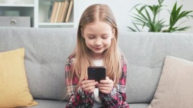 Elinde kablosuz cihazıyla kameraya bakıp gülümseyen sevimli küçük kız. Pozitif mavi gözlü çocuk kişisel akıllı telefondan komik videolar izliyor ve evde vakit geçiriyor..