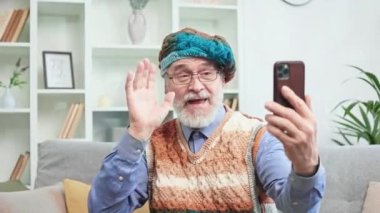 Renkli şapkalı neşeli yaşlı adam evde kişisel akıllı telefonuyla görüntülü konuşma yapıyor. Neşeli büyükbaba komik başlık takıp el sallıyor. Eski nesil ve teknoloji kavramı.