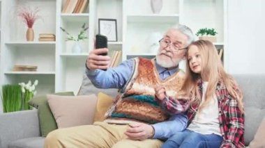 Tasasız büyükbaba ve torun akıllı telefondan selfie çekip eğleniyorlar. Evde vakit geçirirken modern teknolojiyi kullanan aktif, neşeli bir emekli ve küçük bir kız. İki kuşak kavramı.