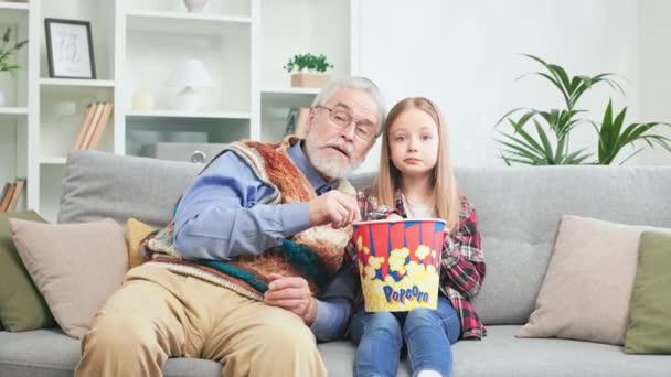 一个好奇的女孩 祖父被有趣的漫画迷住了 吃着美味的爆米花 可爱的孩子们屏住呼吸看电视 祖父在评论事件 — 图库视频影像