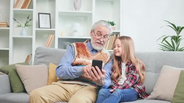 かわいい孫娘と携帯電話でアニメビデオを見て引退した喜びの男 幸せな女の子は祖父の世話をして暇な時間を過ごす 二世代と技術の概念 — ストック動画