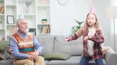 Huzursuz, hareketli bir kız, keyifli bir şekilde oturma odasında dans ediyor. Gri sakallı, son sınıf öğrencisi, pozitif enerji yakalıyor ve torunuyla eğleniyor. Doğum günü kutlaması ve mutluluk kavramı.