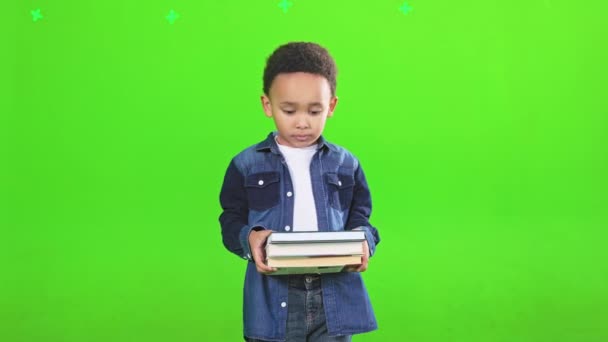 スタジオに立っている間 小さな男の子の本の山を保持笑みを浮かべて 緑の背景に隔離された教科書を持ったデニムシャツを着た愛らしいアフリカ系アメリカ人生徒の正面図 研究概念 — ストック動画