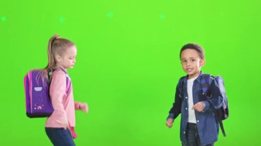 Dans eden ve birlikte atlayan çok ırklı çocuklar ders çalışmaya hazırlar. Mezuniyetin tadını çıkaran, yeşil arka planda izole edilmiş sırt çantalı kız ve oğlanın yan görüntüsü. Okul kavramı, tatiller..
