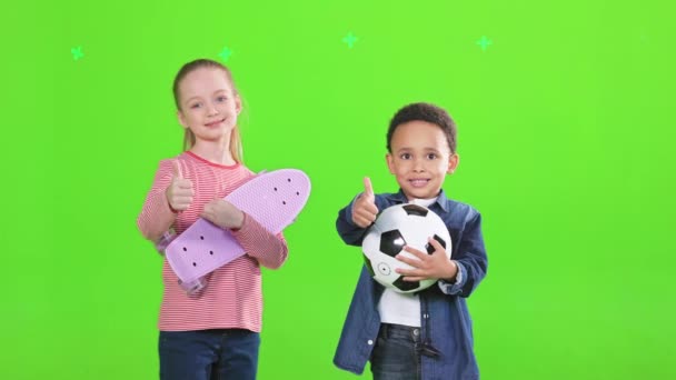 快乐的男孩和女孩与最喜欢的玩具在工作室摆姿势 看到快乐的多种族孩子穿着休闲装 露出大拇指 同时对着相机微笑 被绿色隔离 娱乐的概念 — 图库视频影像