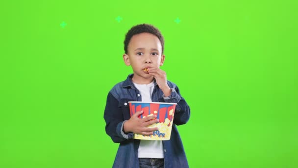 漂亮的非洲裔美国男孩一边在工作室里拿着大爆米花桶一边吃零食 眼前是一个漂亮的小男孩用薯片塞住嘴 动弹不得 与世隔绝 闲暇时间的概念 — 图库视频影像