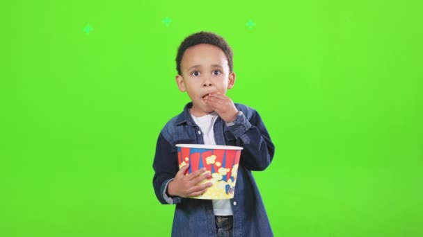 专注于非洲裔美国男孩吃零食 而在工作室里看相机 前看可爱的男孩与爆米花盒咀嚼 思考的东西 孤立在绿色 电影概念 — 图库视频影像