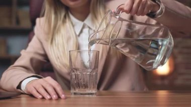 Resmi giysiler içinde, tahta ofis masasının üzerine temiz su döken sarışın kadını yakından çek. Şık bir kadın susuzluğunu hissediyor ve ferahlatıcı bir içecek hazırlıyor..