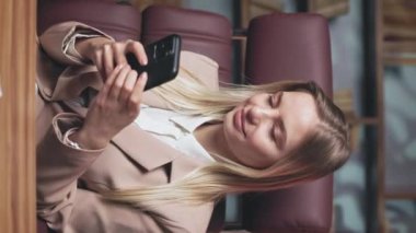 Akıllı telefondan mesaj atan ve kameraya gülümseyen hoş bir genç bayan. Havalı bej ceket giymiş şık ofis sandalyesinde sallanan neşeli sarışın kadın işçi. Dikey video.