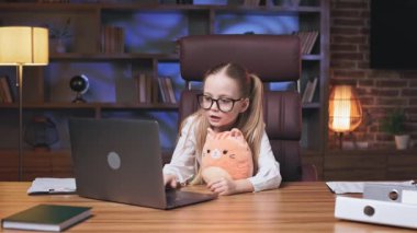 Şirin küçük prenses annesinin ofisinde yumuşak pofuduk oyuncaklarla oynuyor. At kuyruklu çekici bir çocuk kedi yavrusunu pohpohlamak için iş yerini gösteriyor ve ikisi de laptop ekranına bakıyor..