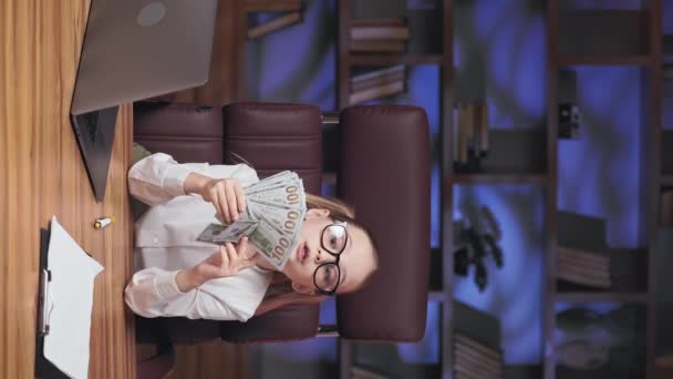 金发碧眼的可爱女孩用美元做扇子 闭着眼睛在面前挥金如土 戴着眼镜 戴着便携设备坐在办公室工作台上的可爱孩子的垂直视频 — 图库视频影像