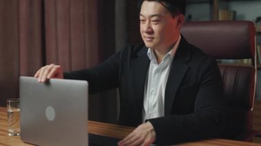 Neşeli Asyalı iş adamının dizüstü bilgisayarı açması ve yeni iş gününe mükemmel bir ruh haliyle başlaması. Çevrimiçi grup sohbetinde arkadaşça selamlaşmalar ve gülümsemeler..