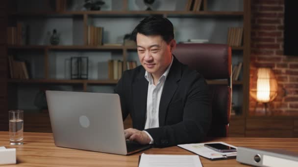 成功的笑着的亚洲人在办公室里与笔记本电脑一起工作 并在短短的休息时间与队友进行在线群聊 正绅士坐在舒适的行政椅上打字 — 图库视频影像