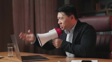 Sinirli şirket patronunun dizüstü bilgisayarda video görüşmesi yapması ve kötü iş performansından dolayı meslektaşlarını azarlaması. Öfkeli Asyalı adam megafonla bağırıyor ve duygusal jestler yapıyor..