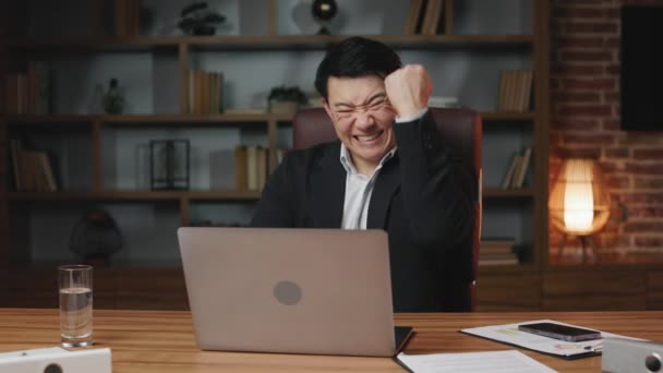 オフィスのローリングチェアに座っている間 仕事の成果を祝い 勝利のジェスチャーを示す興奮した男性ビジネスマネージャー オンライン作業のためのワイヤレスノートパソコンを使用してハンサムなアジアの紳士 — ストック動画