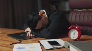 Ahşap ofis masasında uyuyan yorgun finans müdürünün saat 1 'de uyuyakalmaması için ayarlanmış aletleri ve çalar saati. Çok çalışan Asyalı adam dizüstü bilgisayarla uzun süre çalıştıktan sonra dinleniyor..