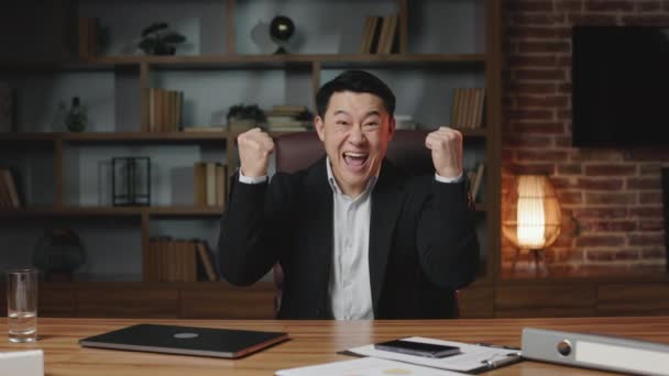 野心的なアジアの従業員は高い給与の仕事の昇進を得 成功を祝う 感情的な男は ワイヤレスガジェットと机の上に座って興奮して拳をきれいにし 勝利のジェスチャーを示す — ストック動画