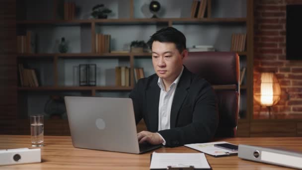 オフィスのデスクトップにワイヤレスコンピュータで座っている間 カメラにポーズをとるアジアの管理者を動機付けました ラップトップを使用したビジネスプロジェクトで働く黒のスーツの深刻な男性大人 — ストック動画