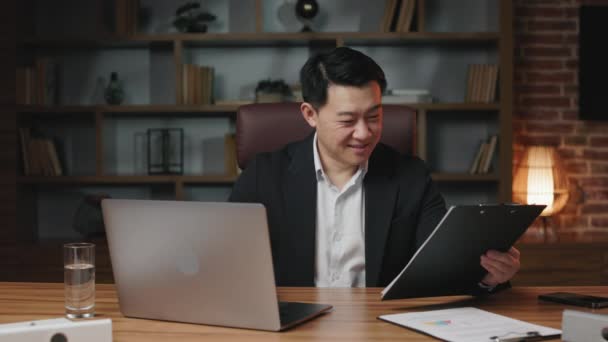 カリスマ的なアジアのビジネスマンは 財務報告書を手に取り コンピュータデータに従って情報を検証します 現代のオフィスで働く正式に服を着た笑顔の男 — ストック動画