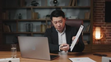 Çevrimiçi personel brifingine katılan Asyalı yönetici düşük indekslerle ilgili endişelerini dile getiriyor. Endişeli adam elinde kalemle kağıtlara bakıyor ve kar düşüşünden bahsediyor..