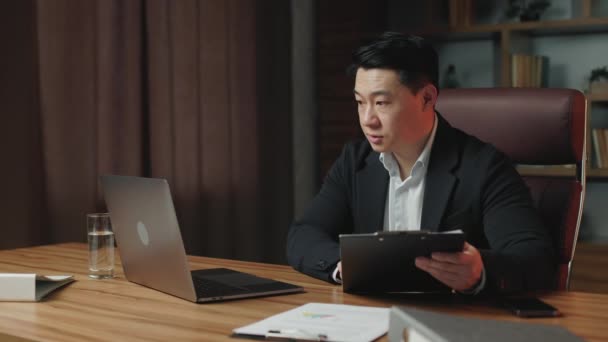 現代的なオフィス空間に滞在しながら ビデオ通話で従業員と話すビジネス服の深刻な会社のCeo ポータブルコンピュータを使用して企業の問題を議論するアジアの起業家 — ストック動画