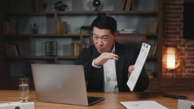 Confident businessman looking and speaking through webcam with teammates while making video conference on laptop at work. Asian supervisor demonstrating papers with graphs and analyzing profit growth.