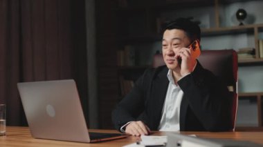 Angle view of self-confident asian real estate agent sitting in modern office with personal computer and talking on cellphone with customer. Cheerful man in stylish black suit making offers for sale.