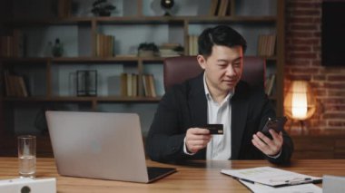 Black-haired company leader sitting at office desktop with wireless computer and making online payment on cellphone. Middle-aged asian man buying things in internet using unlimited credit card.