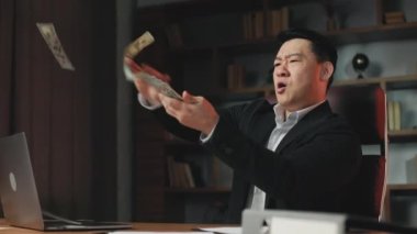 Angle view of prosperous company ceo having fun in own workplace and scattering dollar bills. Thrilled asian man wearing official stylish suit boasting wealthy life.