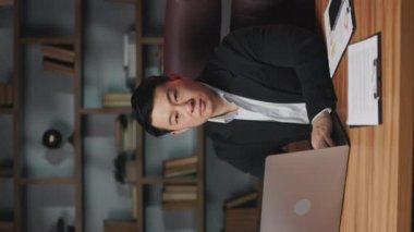 Modern ofisteki yazı masasında portatif bilgisayarla çalışan Asyalı bir adamın dikey videosu. Verimli girişimci kablosuz teknolojiler yoluyla e-ticaret yatırım stratejisi planlıyor.