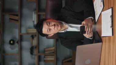 Modern ofiste masa başında otururken elinde cep telefonuyla kameraya gülümseyen başarılı bir iş sahibi. İşte elektronik iletişimi sürdüren etkili Asyalı yöneticinin dikey videosu.