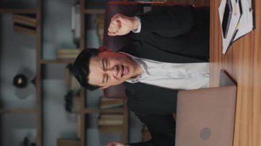 Bilgisayarın arkasında ofis sandalyesinde oturmuş zafer dansı yapan heyecanlı orta yaşlı bir çalışan. Neşeli Asyalı profesyonel büyük anlaşmayı kutlarken içsel ritmiyle sallanıyor. Dikey video.