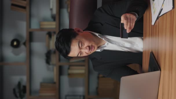 ビジネスススーツの感情的な労働者は コンピュータとオフィスデスクで喜んでクレジットカードで腕を上げます 仕事で電子バンキングを使用して長い待ち望まれたオンライン購入を作るアジア人男性の垂直ビデオ — ストック動画