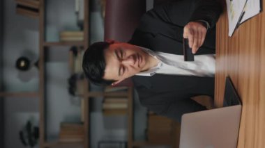 Ofisteki banka kartına bakarken portatif bilgisayarda daktilo yazan dikkatli bir girişimci. Odaklanmış Asyalı finansör plastikleri kontrol ediyor. Dikey video.