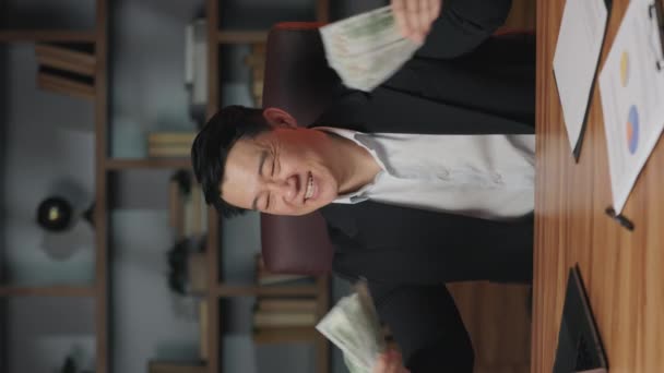 オフィスの椅子に座っている間 ファンを振る正式な服で成功したアジア人男性の垂直ビデオ 仕事で現金で楽しんでいる間 仲間の収入に関わる幸せな豊かなビジネスオーナー — ストック動画