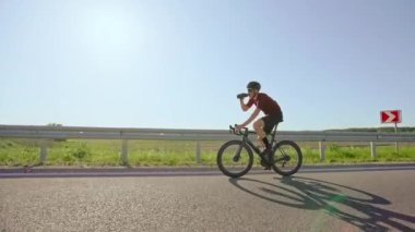 Asfalt yolda bisiklet sürerken spor şişesinden içki içen profesyonel bisikletçinin yan görüntüsü. Dayanıklılık eğitimli sporcu yaz günü yüksek yoğunluklu idman gezisinde susuz kalmıyor.