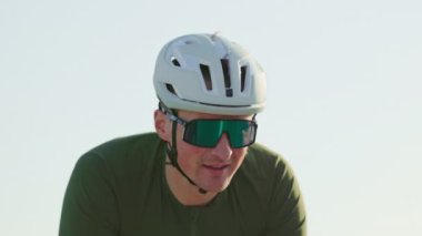 Spor kıyafetli çekici bir erkeğin yaz günbatımında yarış bisikleti eyerinden inişini yakından izleyin. Güç egzersizi sırasında tepeye çıkarken pedalların üzerinde duran beyaz bir sporcu..